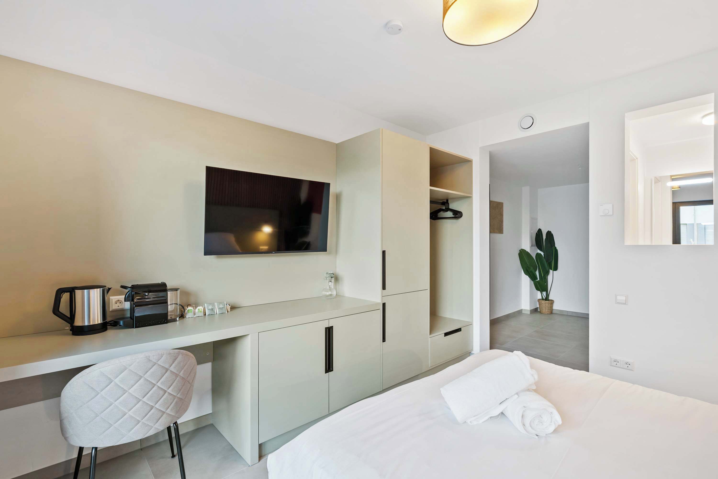 Una habitación de hotel moderna con una cama, un escritorio con silla, televisor, cafetera y una planta en maceta.