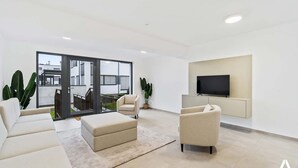 A modern living room with a flat-screen TV, two sofas, a coffee table, and a large sliding glass door leading to an outdoor balcony.