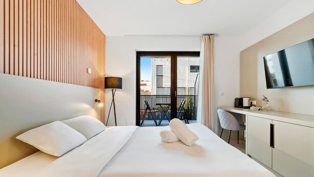 A modern hotel room with a large bed, a desk, a flat-screen TV, and a balcony with a view of the city.