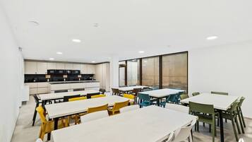 Un comedor moderno con mesas y sillas blancas, una cocina al fondo y ventanas amplias.