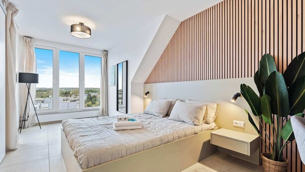 Un dormitorio moderno con una cama grande, mesitas de noche, una lámpara de pie, una planta en maceta y vistas amplias a través de grandes ventanales.