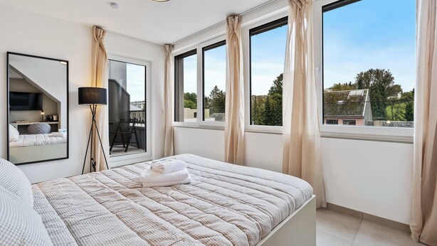 Ein Schlafzimmer mit einem großen Bett, einem Spiegel, einer Stehlampe und Blick ins Freie durch große Fenster.
