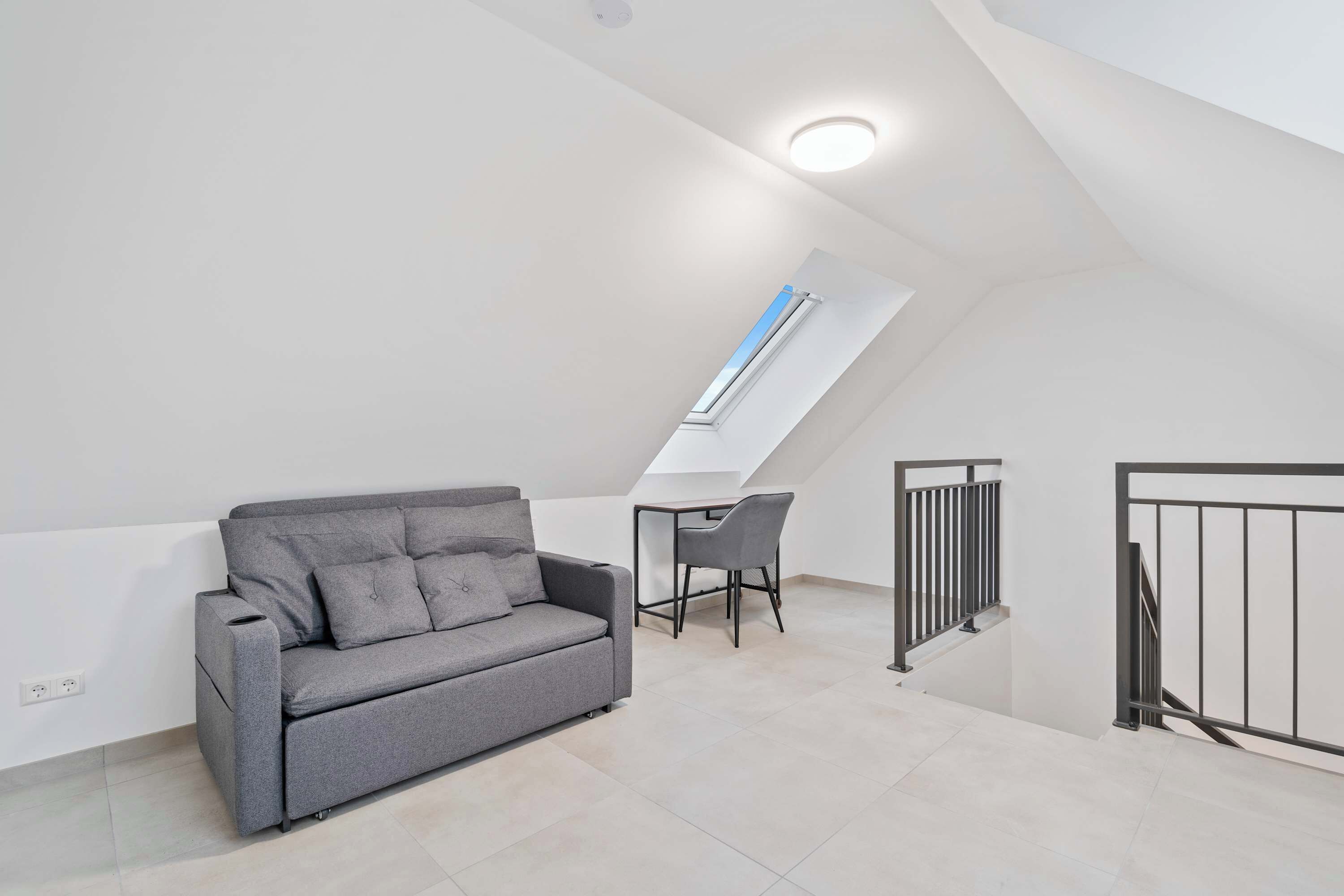 Un dormitorio tipo loft moderno con techo inclinado, un sofá gris, un escritorio pequeño y una silla.