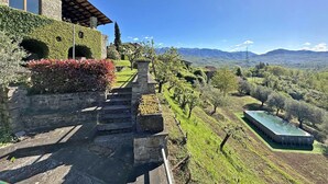 A stone building with a stone staircase leading to a terrace, surrounded by greenery and a view of distant mountains.