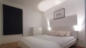 A bedroom with a bed, a bedside table, a wall-mounted light, and a framed word on the wall.
