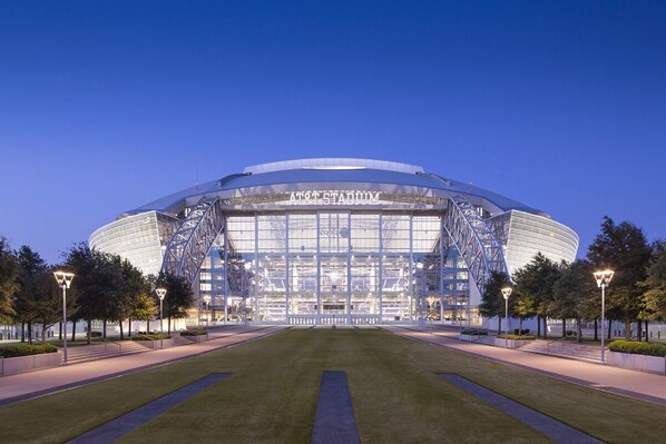 A stadium with a large, curved roof and a glass facade, surrounded by well-maintained lawns and street lamps.