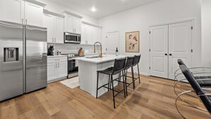 A modern kitchen with white cabinetry, stainless steel appliances, and a central island with bar stools. - Stadium Steps Away | Stylish Townhome w/ Parking (Arlington)