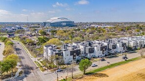 A modern residential area with compact, multi-story buildings, a stadium, and a clear road. - Walk to AT&T Stadium | Luxury Townhome | Parking (Arlington)
