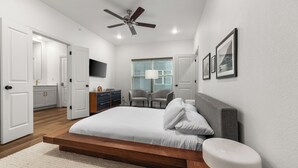 A modern bedroom with a wooden bed, a ceiling fan, a desk with chairs, and a large window with blinds. - Stadium Steps Away | Stylish Townhome w/ Parking (Arlington)