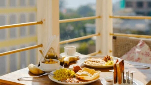 Une table à manger avec différents plats et une tasse, placée devant une fenêtre donnant sur un bâtiment et des montagnes.