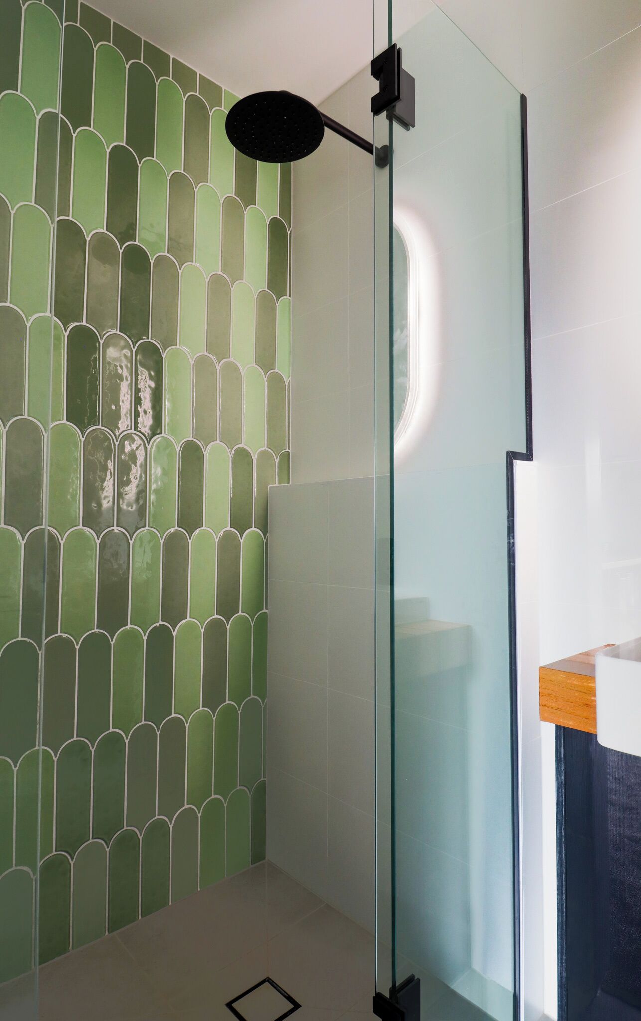 A modern bathroom with a green tiled wall, a glass shower enclosure, and a wooden countertop.