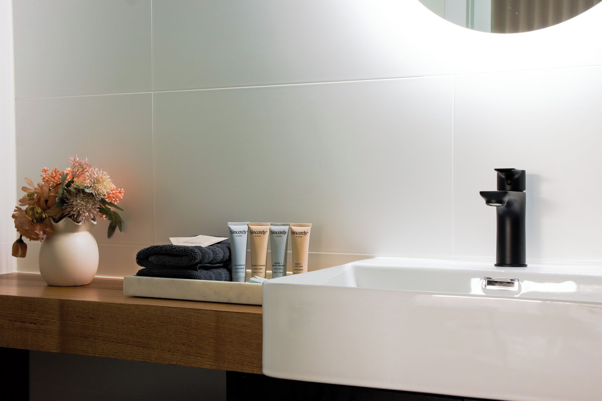 A bathroom with a white sink, black faucet, and toiletries including a vase with flowers, towels, and skincare products.