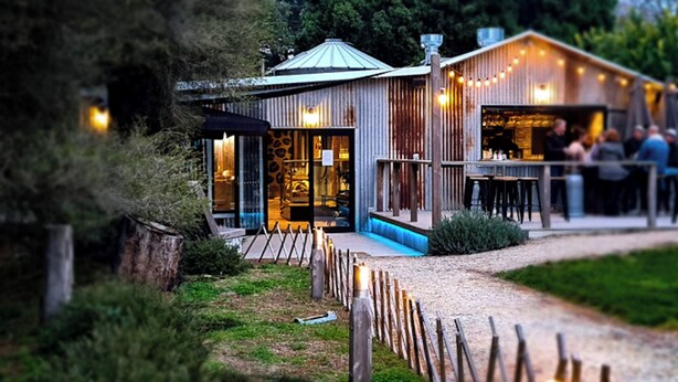 A rustic building with a metal roof, a wooden deck, and a gravel path leading to the entrance.