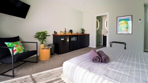 A modern bedroom with a bed, a sofa, a small kitchenette, and a TV mounted on the wall.