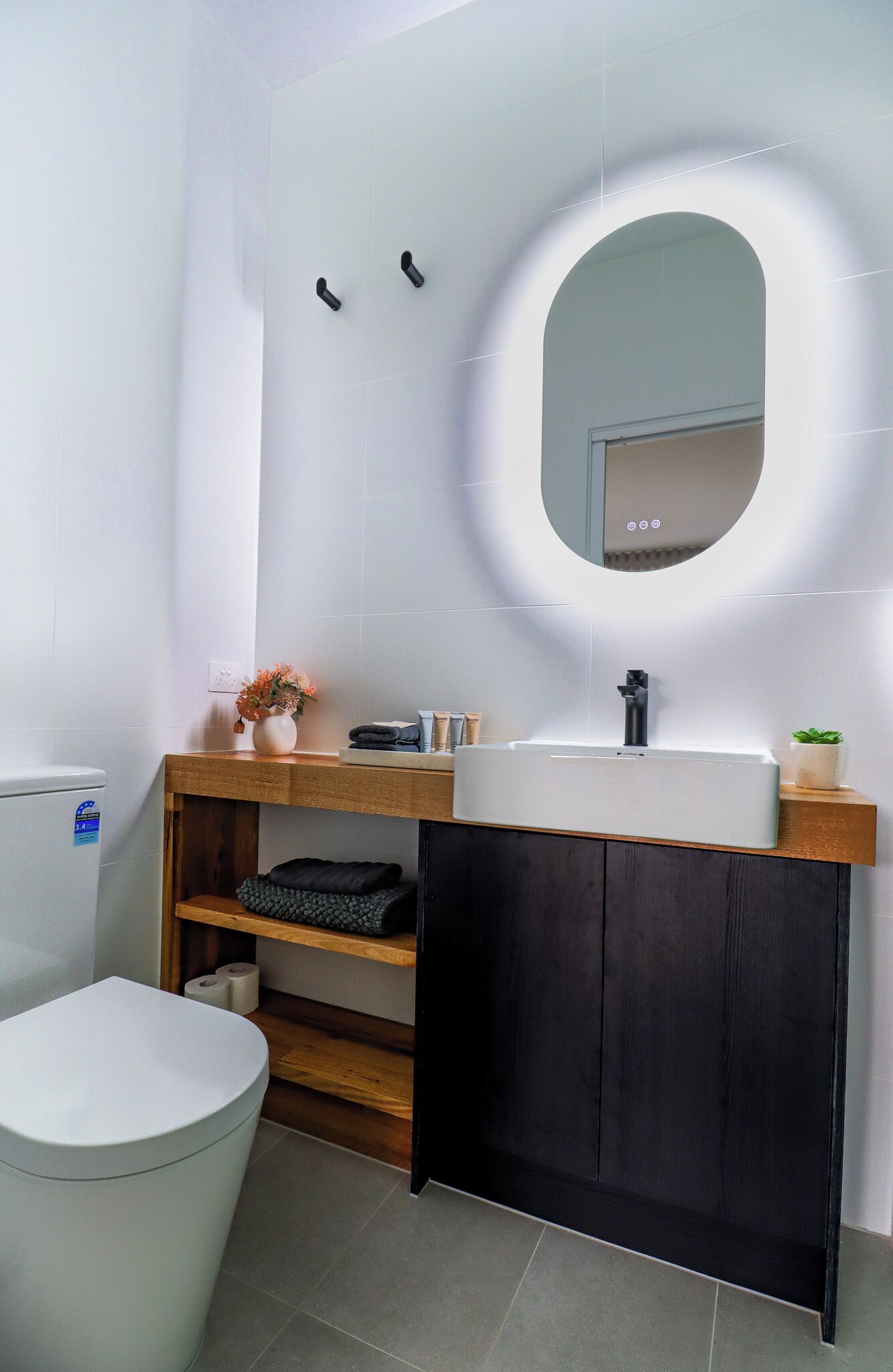 Un baño moderno con inodoro blanco, tocador de madera con mueble negro y espejo con marco iluminado.