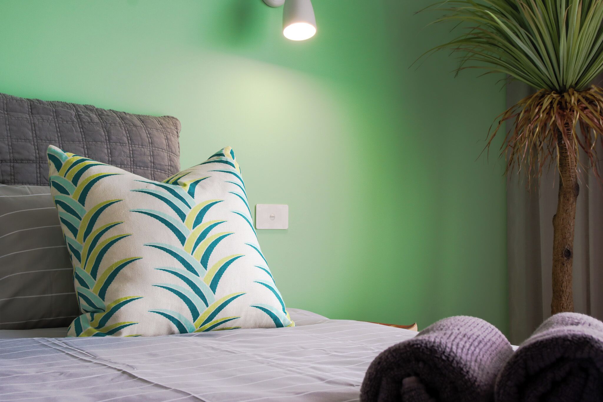 Een bed met een witte spreideken, een kussen met bladprint en een bloempot met plant.