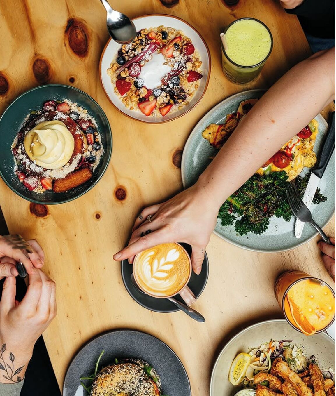 A wooden table with various dishes, including a plate of cereal with fruits, a plate of vegetables, a plate of fish, and a cup of coffee.
