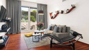 A modern living room with a sofa, a coffee table, and a chair. There is a large window with curtains, a plant, and a view of the outdoors. - Serena Calm Pool House (Vari-Voula-Vouliagmeni)