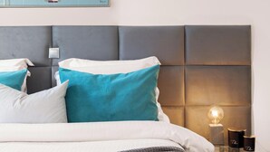 A neatly made bed with a grey headboard, white and teal pillows, and a grey knitted throw. A framed painting of boats is on the wall above the bed. - Serena Calm Pool House (Vari-Voula-Vouliagmeni)