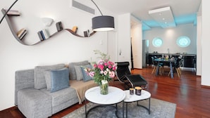 A modern living room with a grey sofa, a coffee table, and a vase of flowers. There is a dining area with a table and chairs in the background. - Serena Calm Pool House (Vari-Voula-Vouliagmeni)