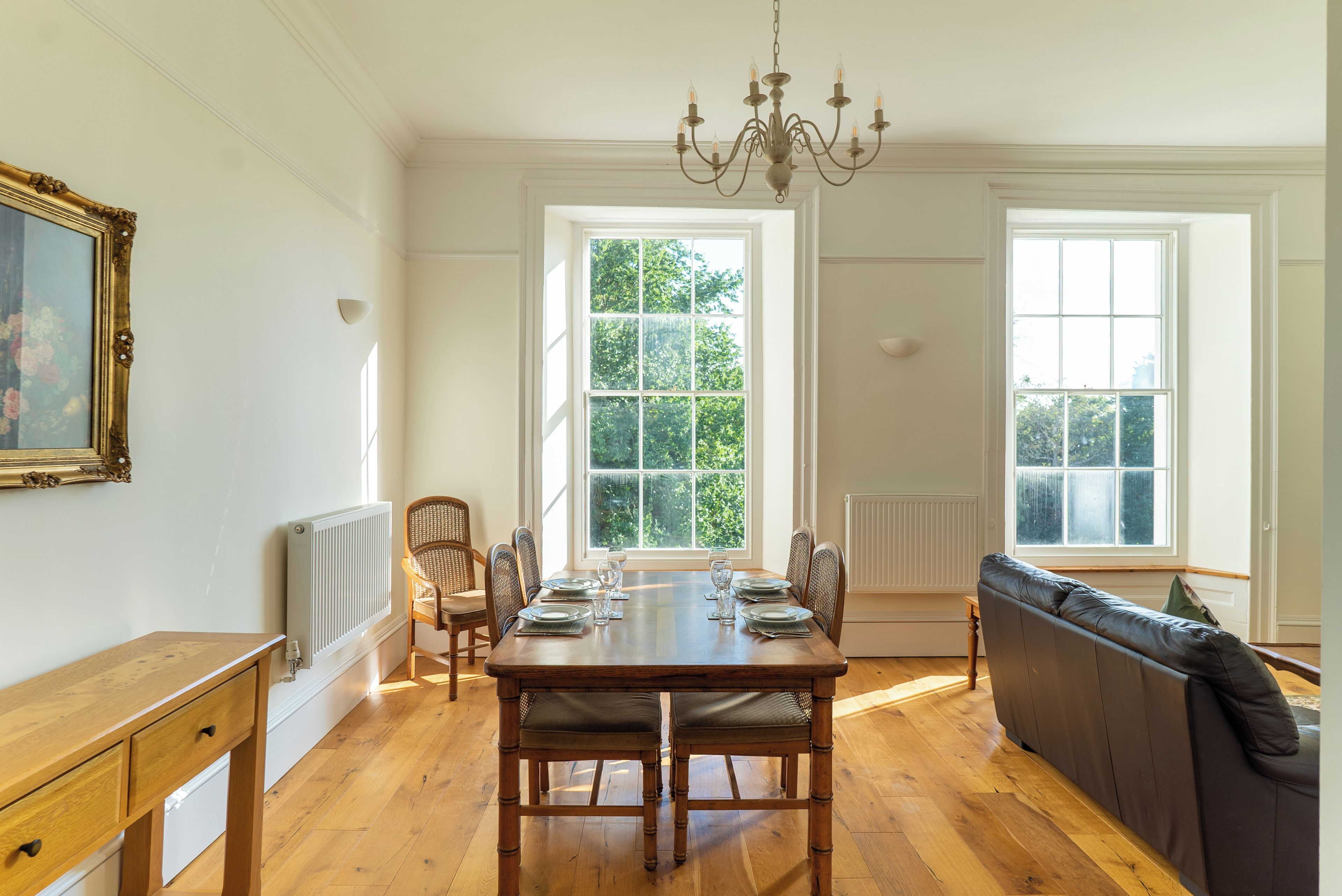 A dining room with a wooden table set for six, surrounded by chairs, a sofa, and a chandelier.