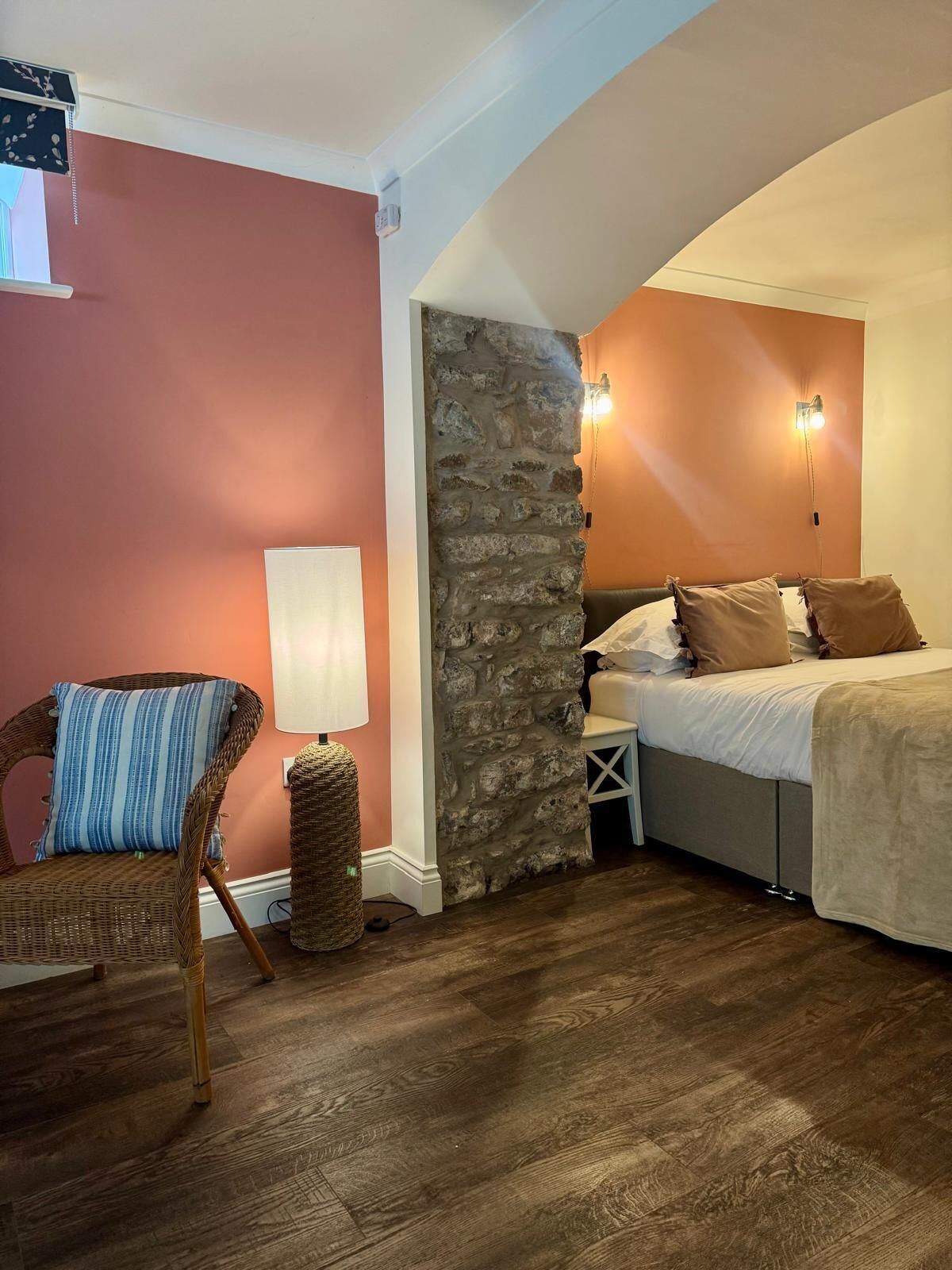 A bedroom with a stone accent wall, a bed with pillows, a wicker chair, and a bedside table with a lamp.