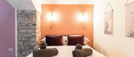 Quarto de hotel com parede de destaque em pedra, uma cama com duas almofadas e duas luzes fixadas na parede.
