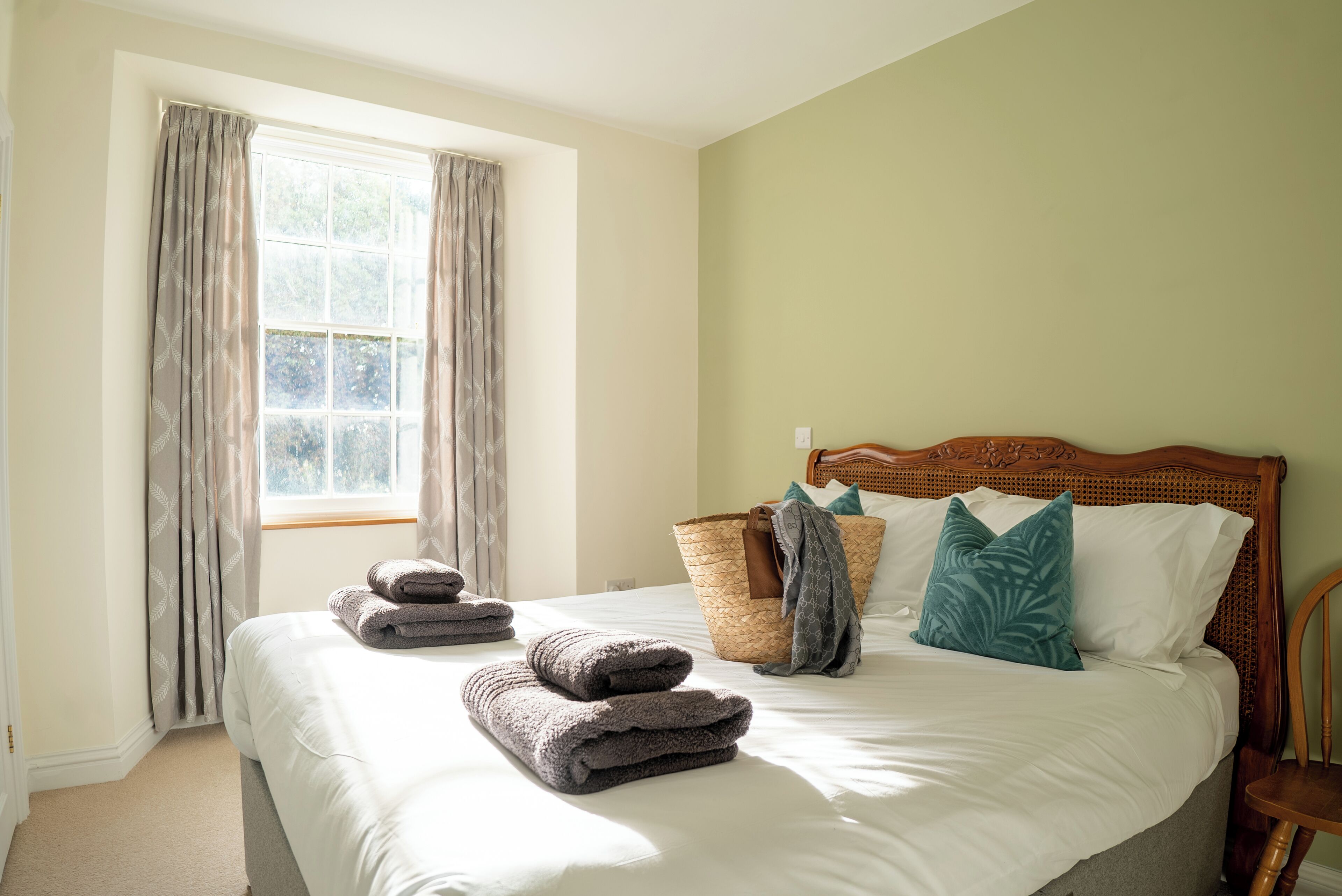 A neatly made bed with folded towels, a wicker basket, and a wooden headboard in a room with a window and a plain wall.