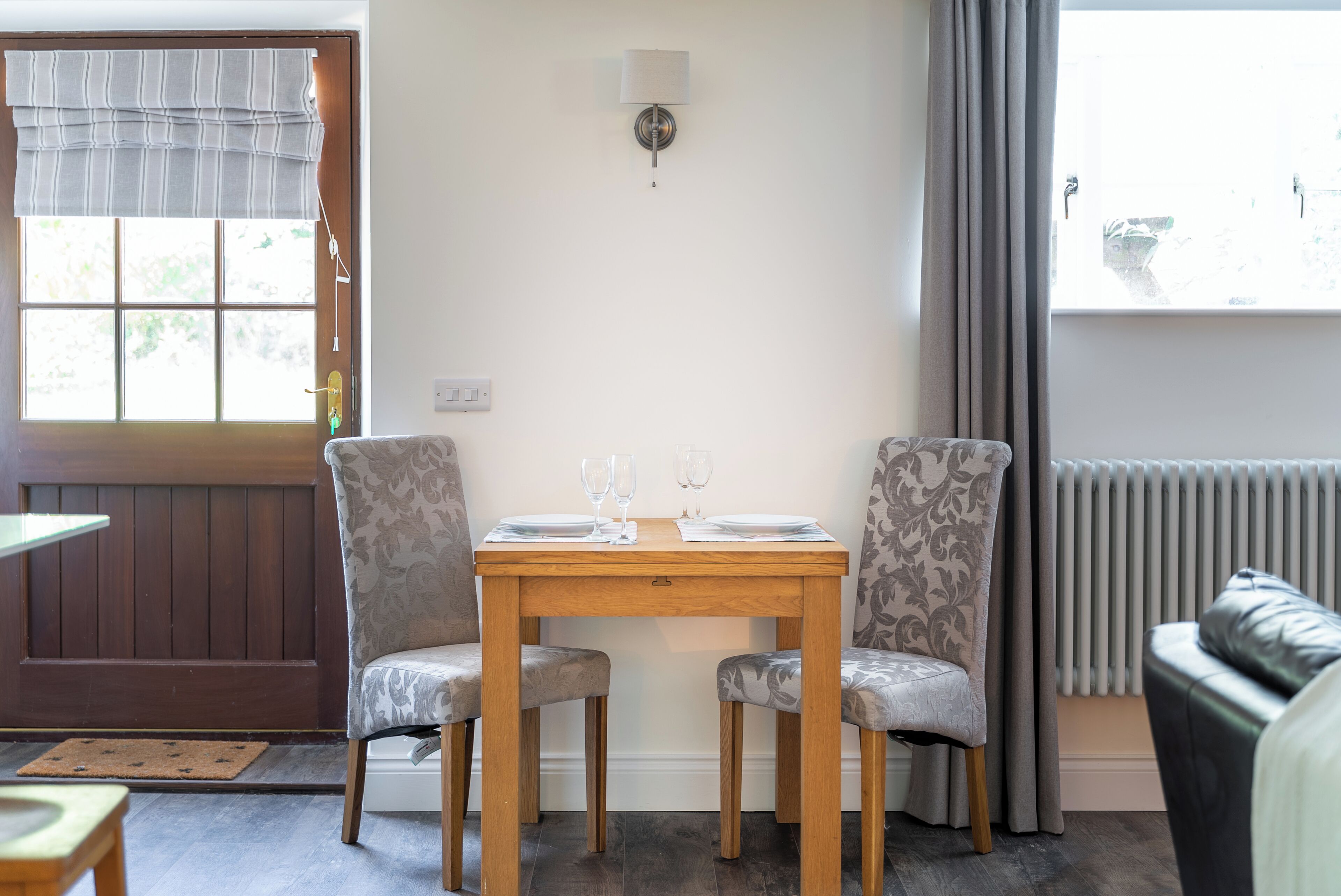 A dining area with a wooden table, two patterned chairs, a wall-mounted light, and a window with curtains.