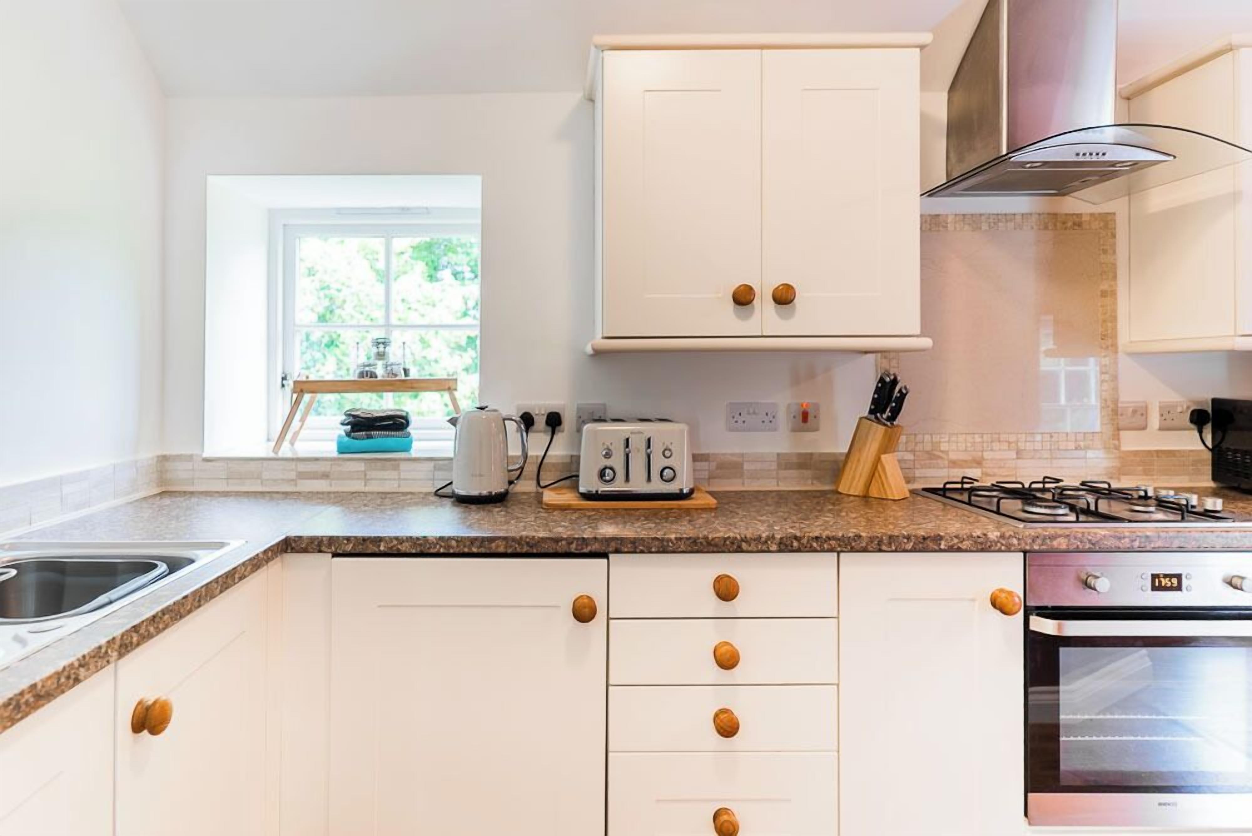 Una cocina moderna con armarios blancos, pomos de madera, estufa a gas, horno empotrado y ventana con vista a la vegetación.