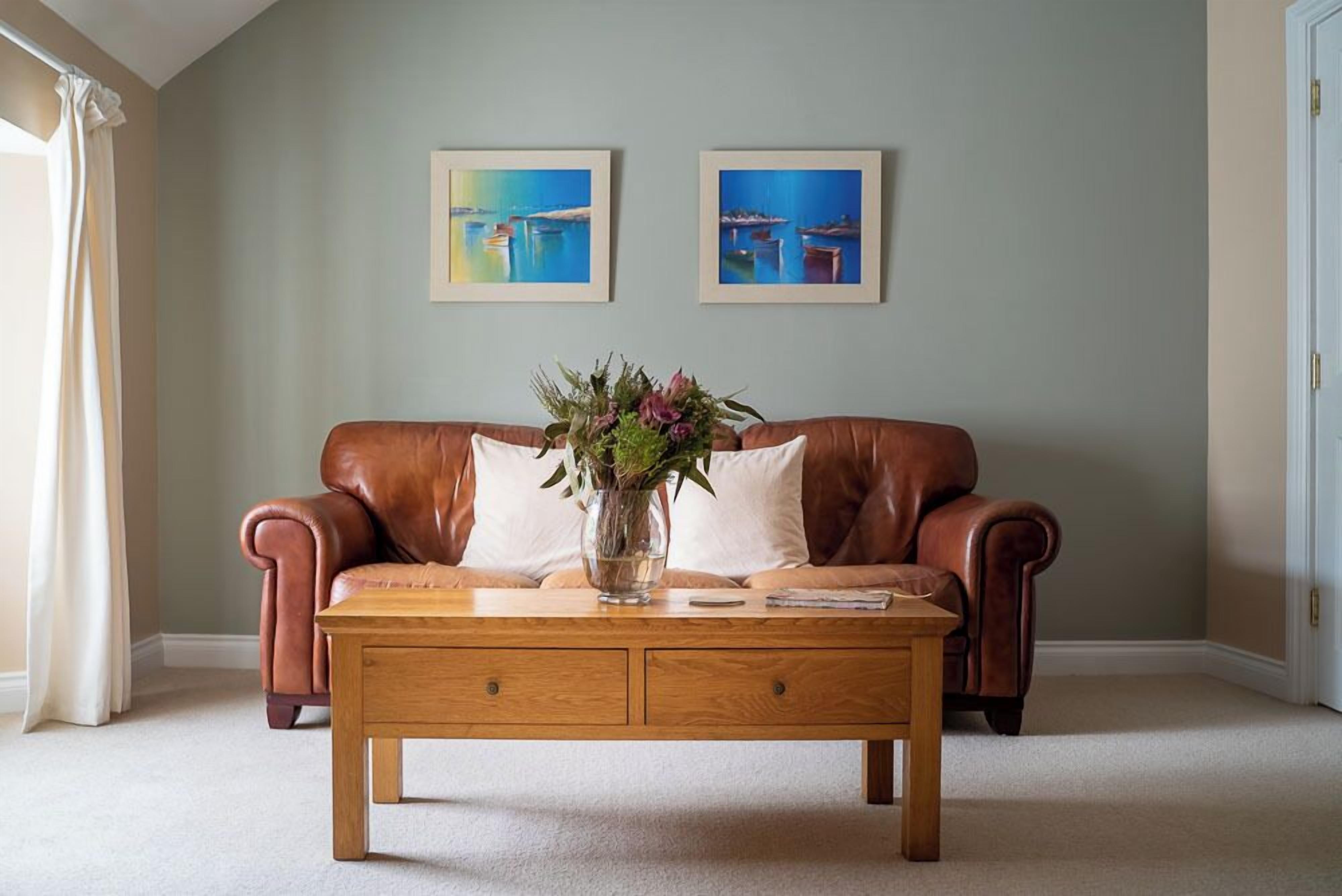 A living room with a brown leather sofa, a wooden coffee table with a vase of flowers, and two framed paintings on the wall.