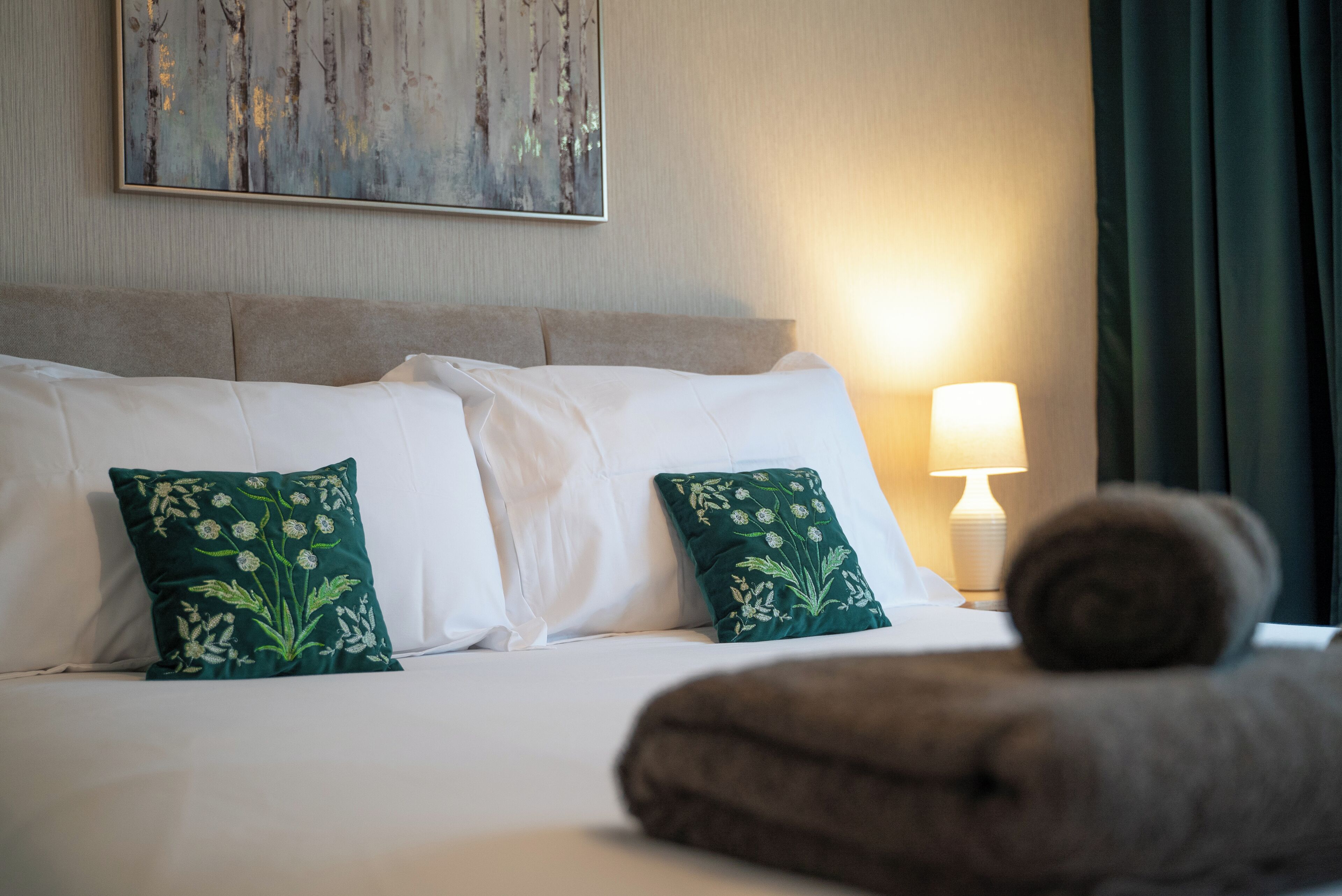 A neatly made bed with white linens and two decorative pillows, a bedside lamp, and a rolled towel on the bed.