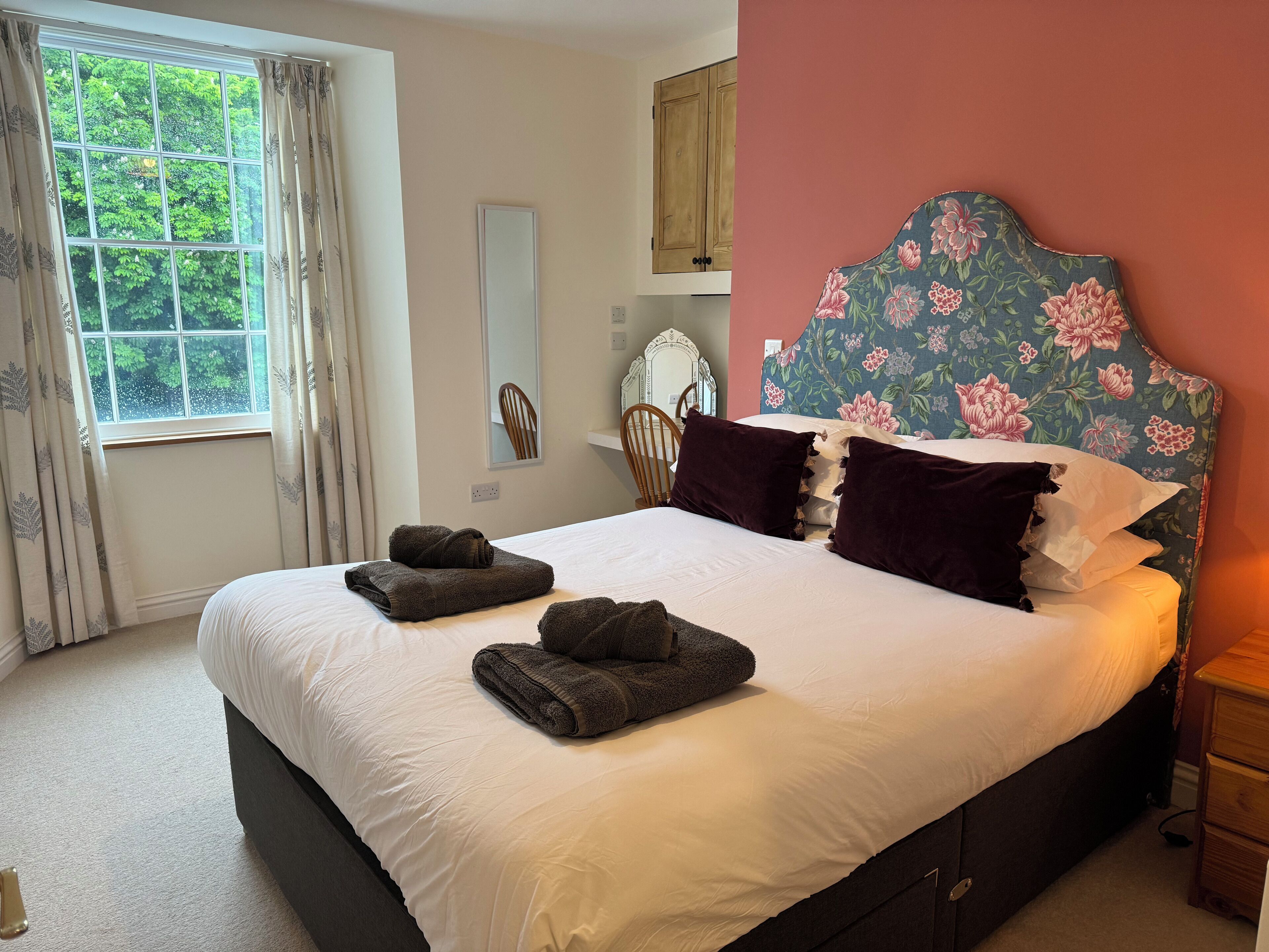 A neatly made bed with folded towels, a wooden headboard with floral patterns, a mirror, and a window with curtains.