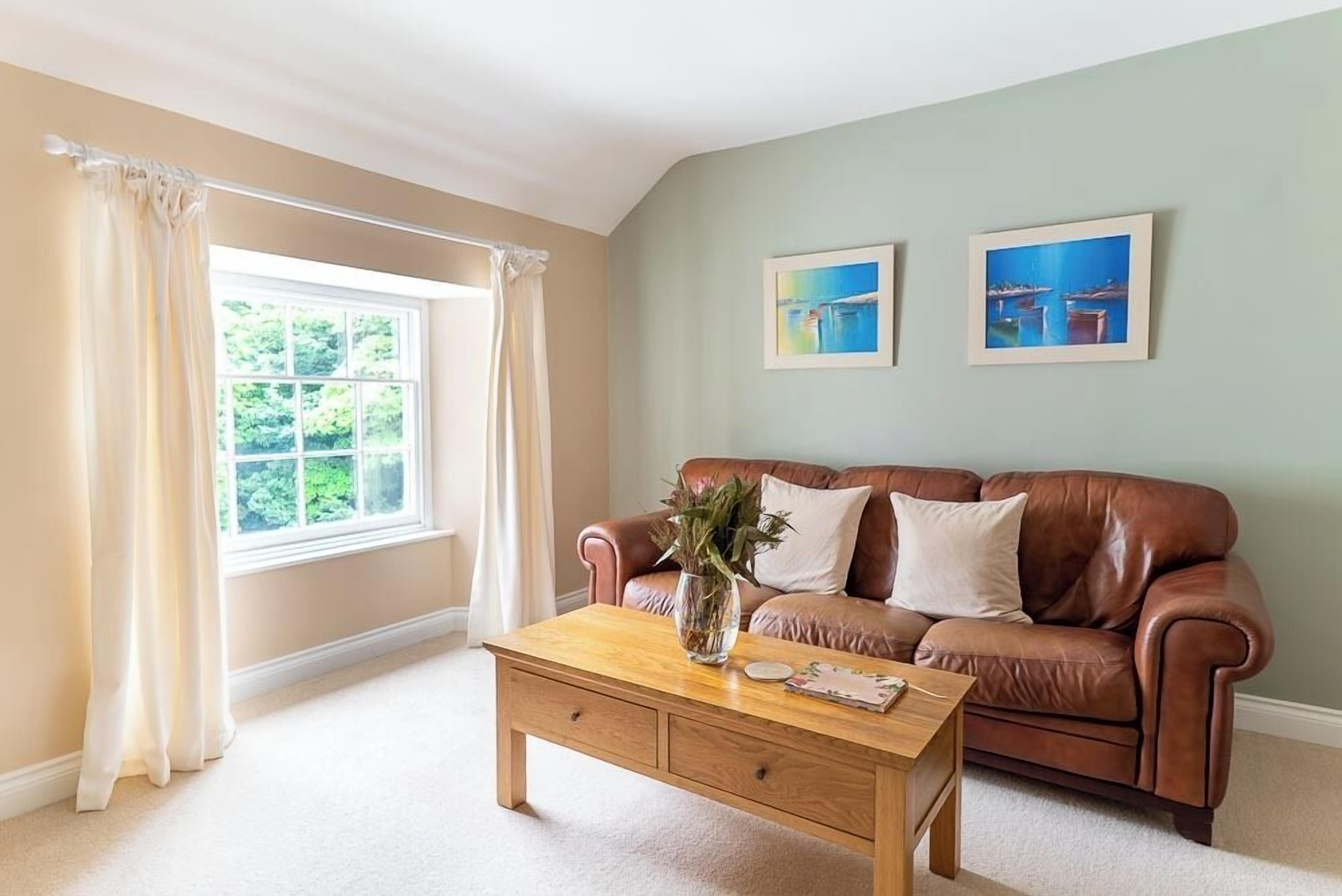 A living room with a brown leather sofa, a wooden coffee table with a vase and a book, and two framed pictures on the wall.