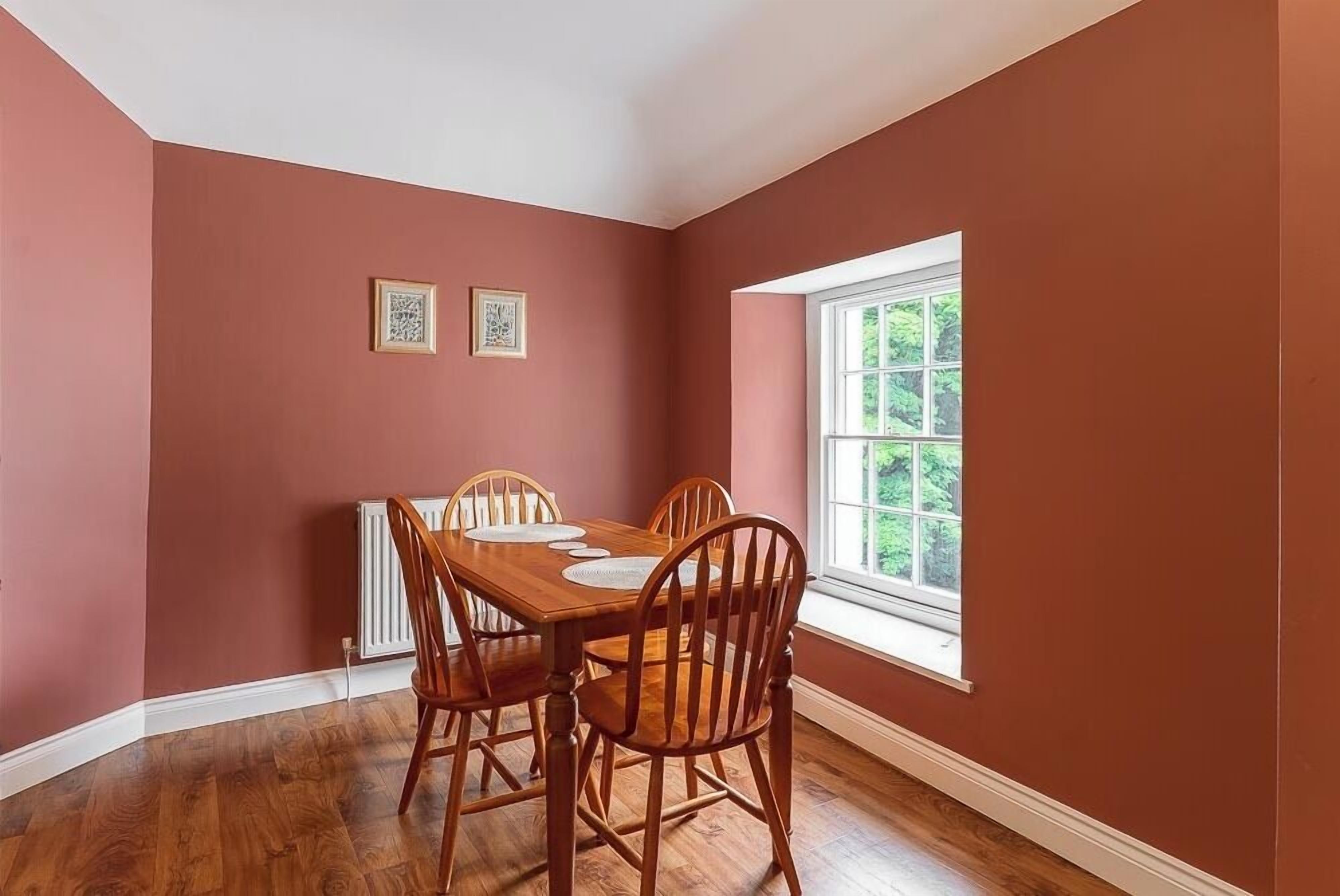 Un comedor con mesa y sillas de madera, una ventana con vista a la vegetación y dos cuadros en la pared.