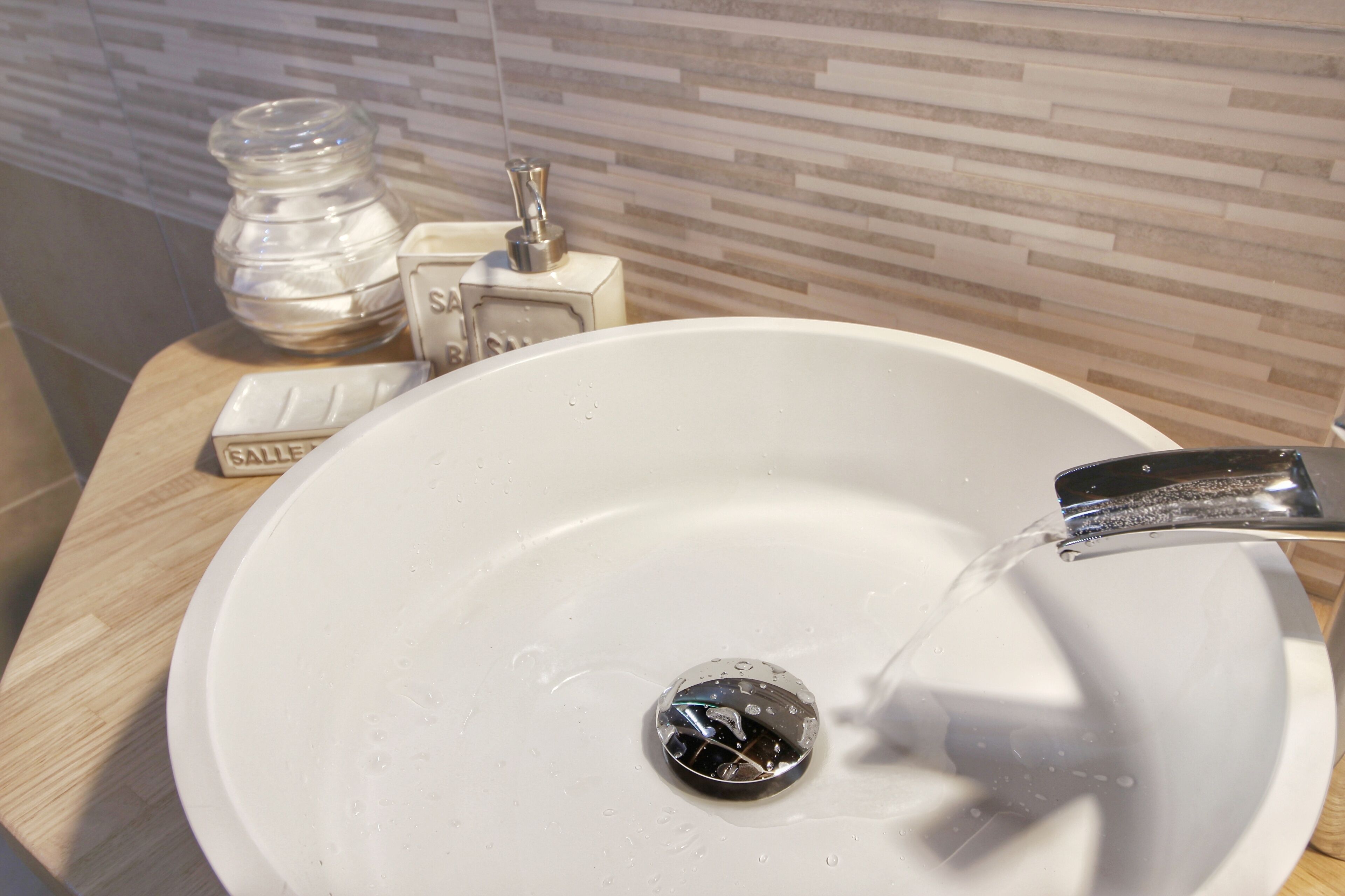 Un lavamanos blanco con agua corriente, un dispensador de jabón y un frasco de vidrio sobre una encimera de madera.