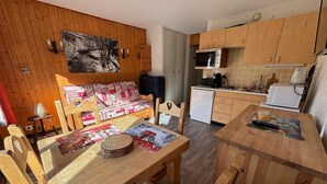 A cozy cabin-style room with a wooden dining table, a sofa with a patterned cover, a small kitchen area with a microwave and refrigerator, and a wooden cabinet.