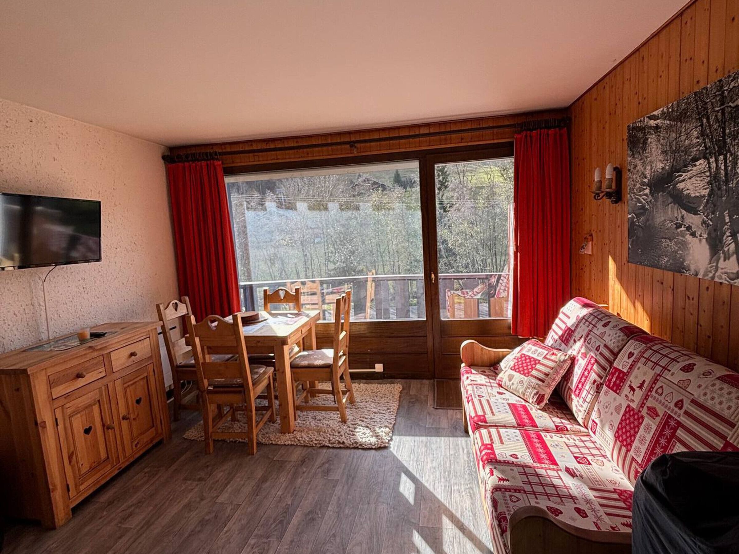 Ett mysigt rum med ett matbord och stolar i trä, en soffa, en tv som är monterad på väggen och ett stort fönster med röda gardiner.