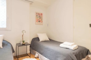 A small, clean room with two beds, a small table with a lamp, and a painting on the wall.