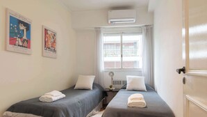 Een kamer met twee bedden, witte handdoeken, een raam met gordijnen, een airconditioner en twee posters aan de muur.