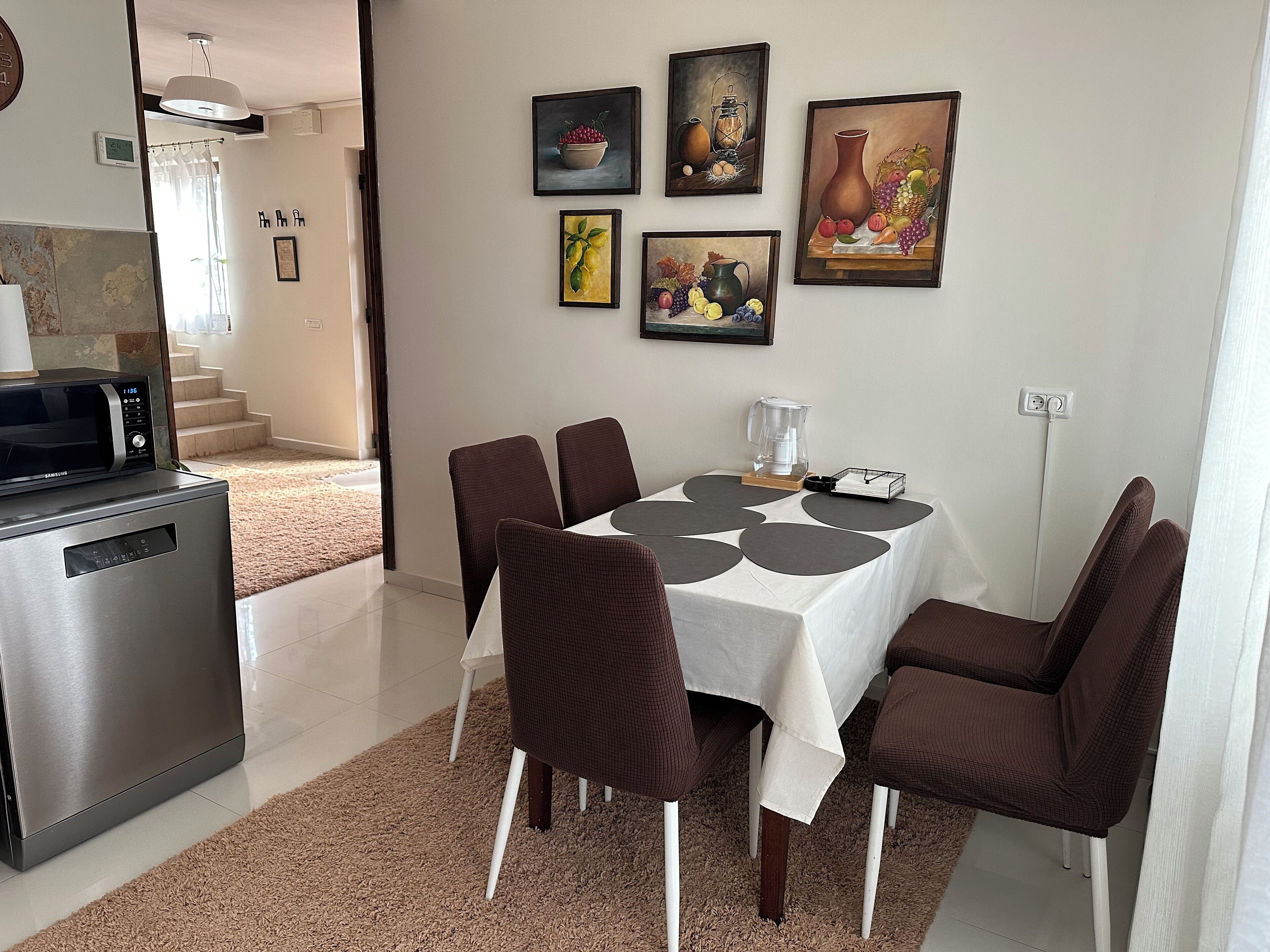 A dining area with a table set for six, surrounded by brown chairs, a coffee maker, and a fruit bowl on the table.