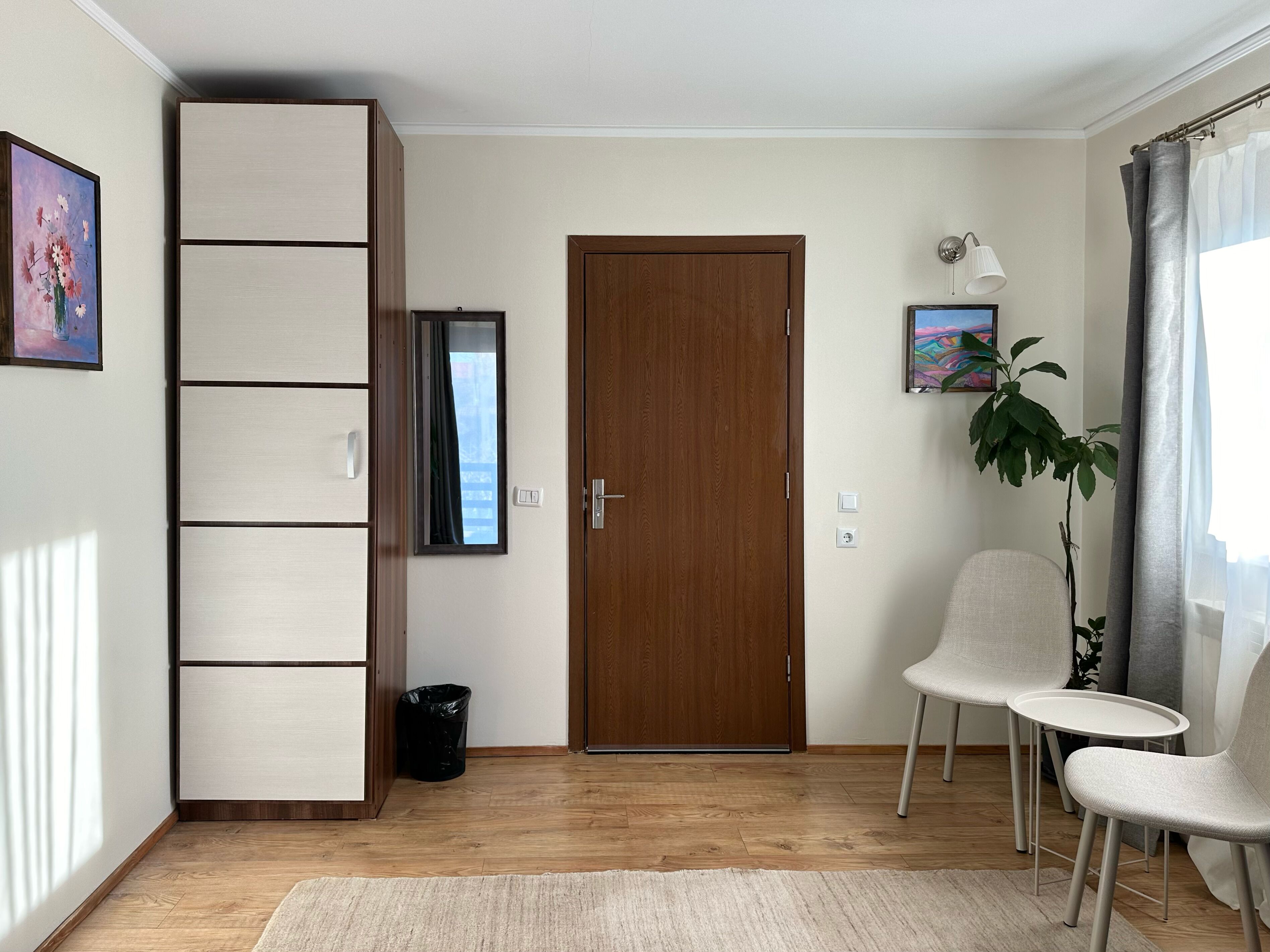 Una stanza con un'armadio in legno, una porta, un tavolino con due sedie, una pianta in vaso e una finestra con tende.