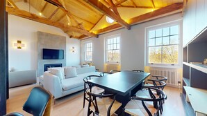 A modern living room with a large table, sofa, and chairs. There is a fireplace, a television mounted on the wall, and large windows providing natural light. - 3-bedroom Penthouse in Knightbridge (London)