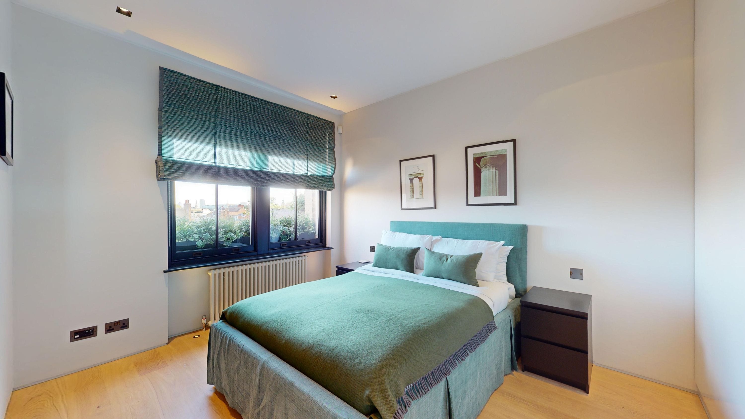 A modern bedroom with a large bed, a window with blinds, a radiator, and two framed pictures on the wall.
