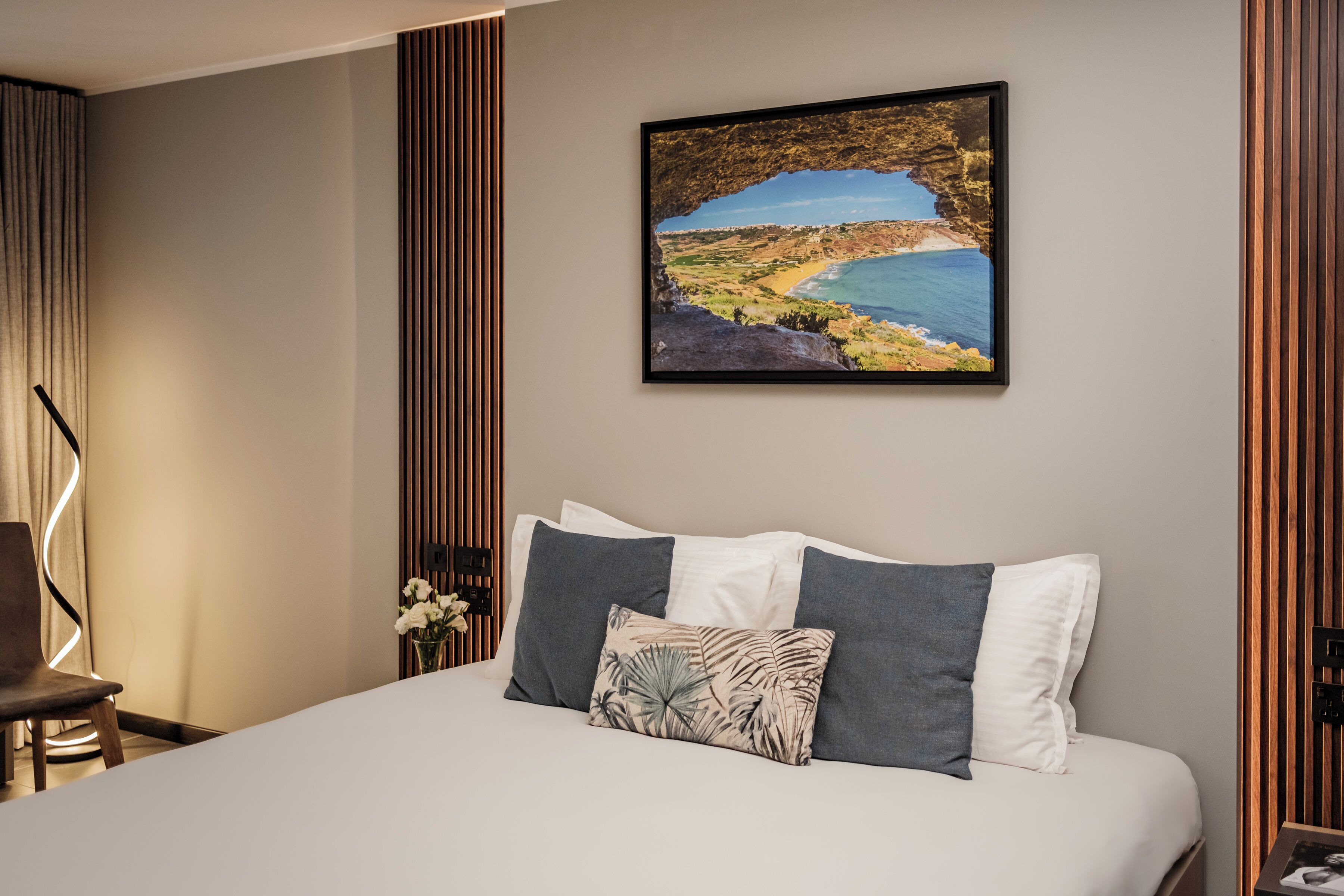 A neatly made bed with a coastal landscape painting above it, a wooden panel wall, and a chair with a light fixture.