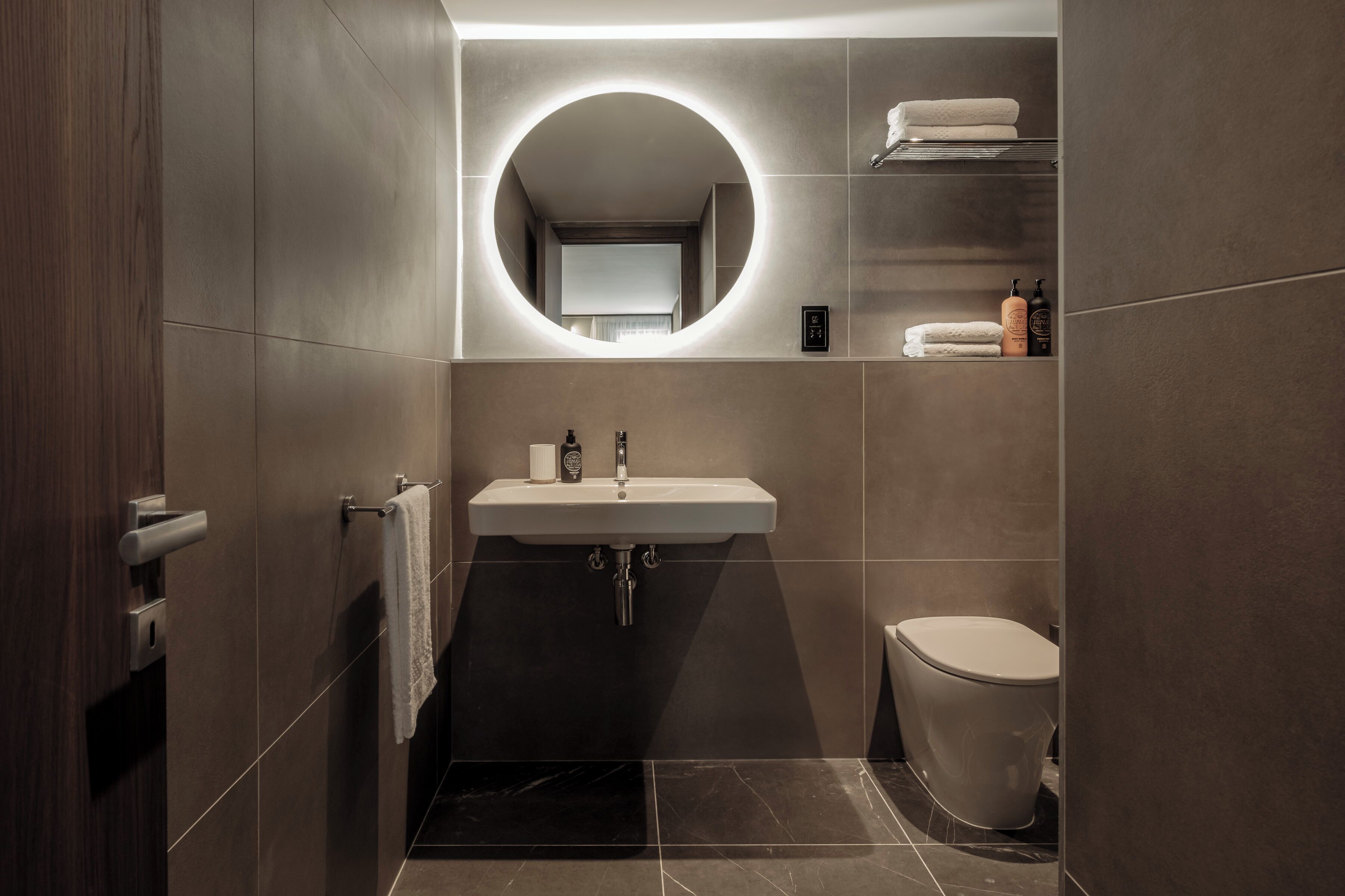 Een moderne badkamer met een ronde spiegel, een wastafel, toilet en een handdoekrek.