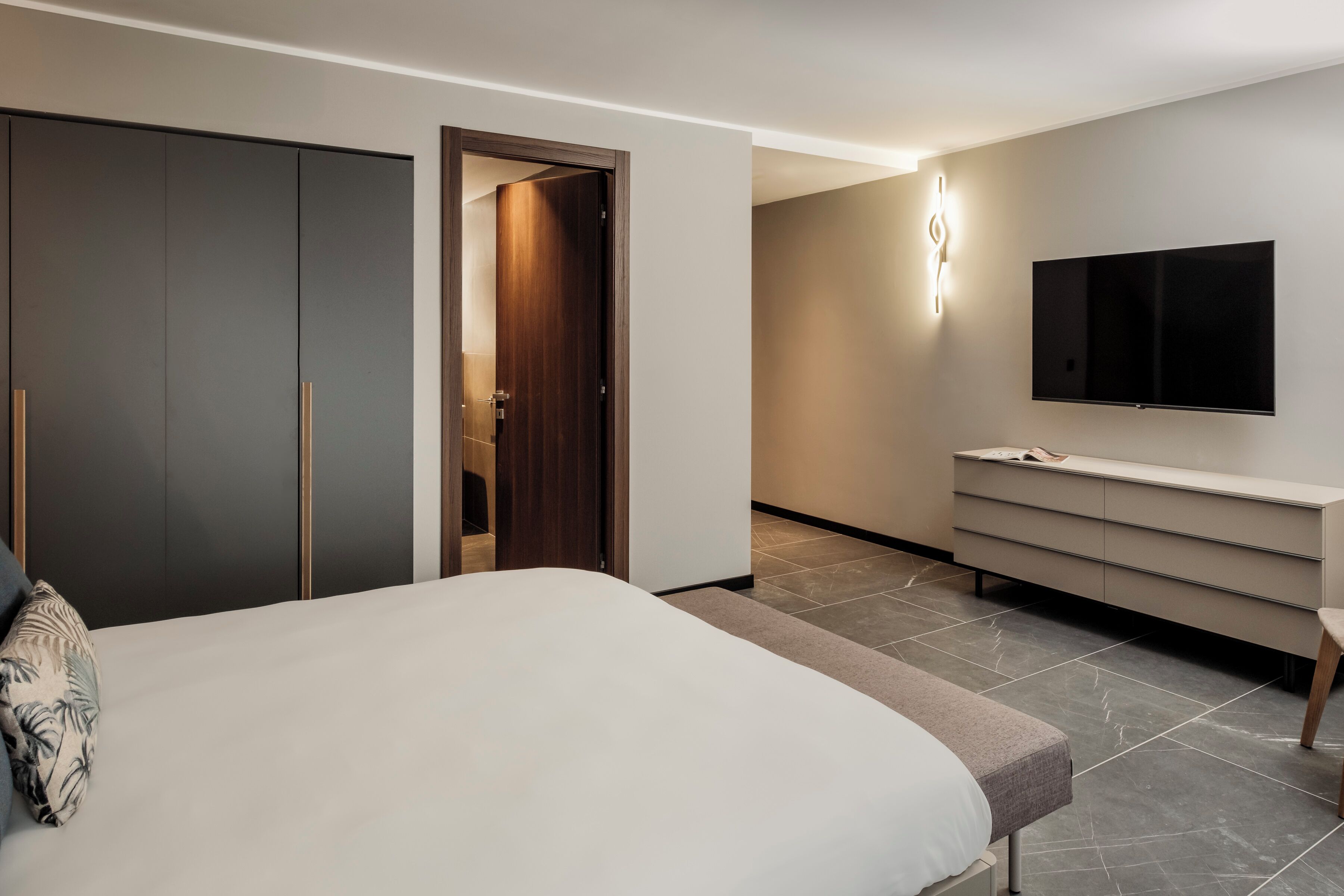 A modern hotel room with a large bed, a wardrobe, a vanity with a mirror, and a flat-screen TV mounted on the wall.