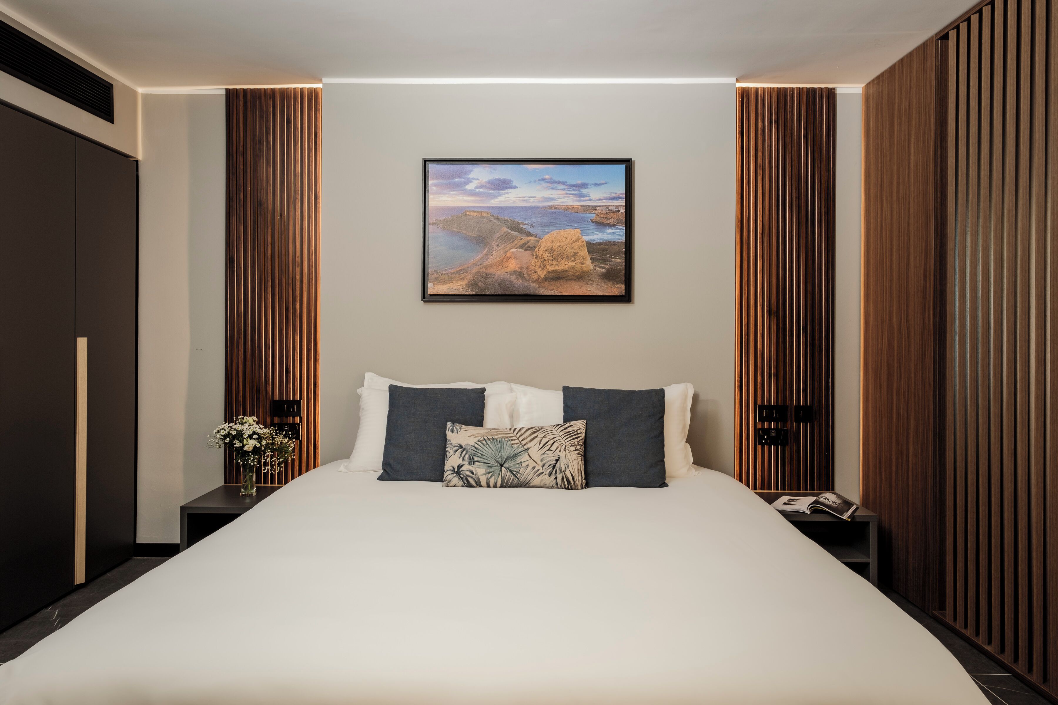 A modern bedroom with a large bed, a nightstand with a vase of flowers, and a framed picture on the wall.