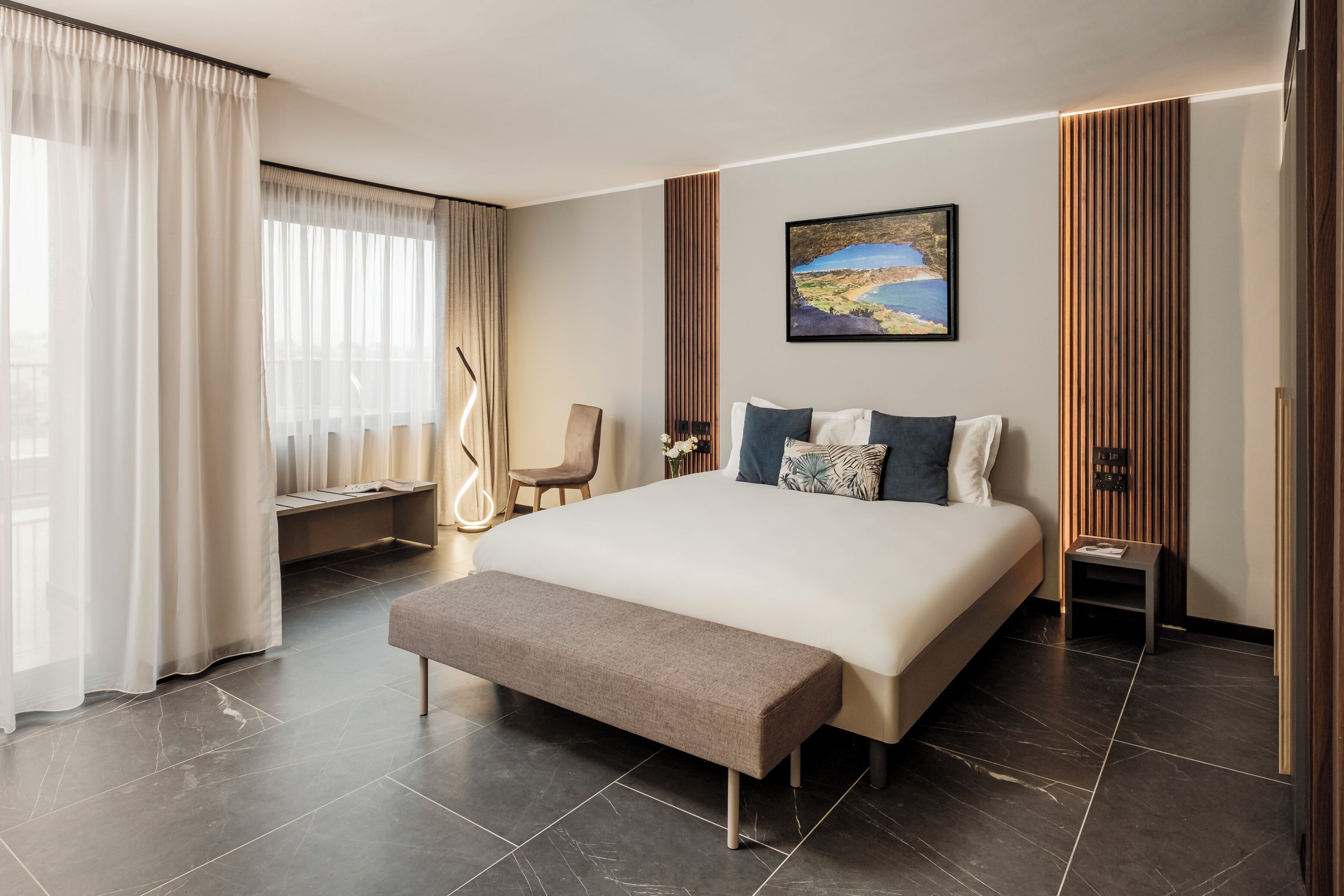 A modern hotel room with a large bed, a bench, a bedside table, and a view of the outdoors.