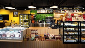 A cafe with a coffee machine, a display of pastries, and a green wall. - Isabella Suites (Gzira)